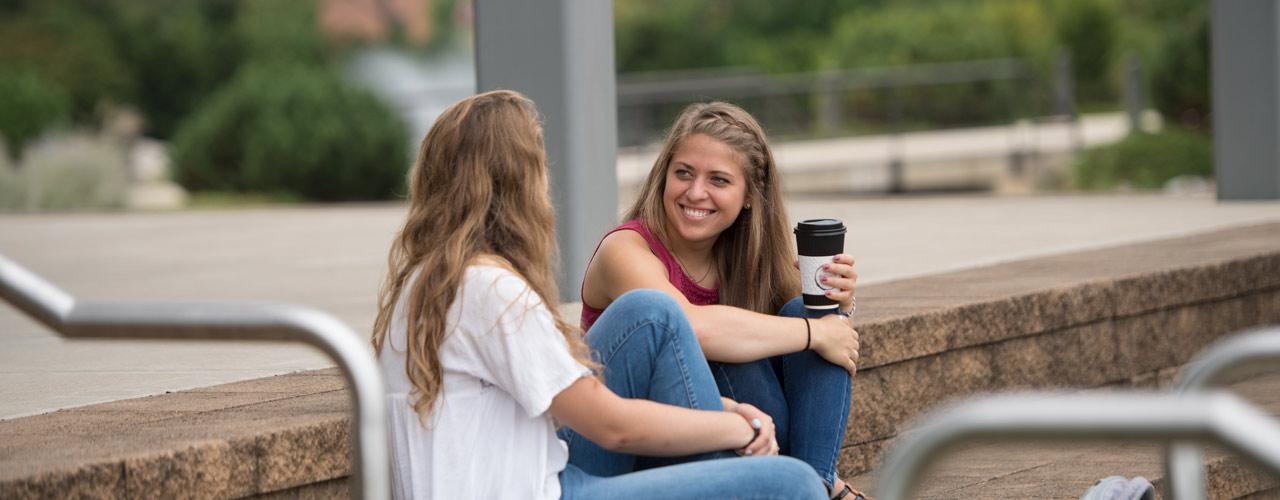 Two female Cedarville students smiling with coffee on stone steps outside of The Center for Biblical and Theological Studies
